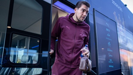 Manchester City player, wearing team training jacket and pants with Puma logo, exits team bus holding a water bottle and a small bag.