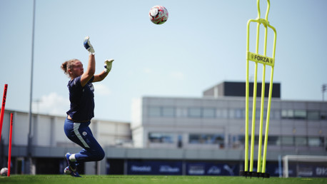 A goalkeeper is in training, wearing blue gear and catching a ball in mid-air on a grass field.