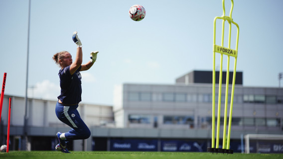 A goalkeeper is in training, wearing blue gear and catching a ball in mid-air on a grass field.