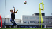 A goalkeeper is in training, wearing blue gear and catching a ball in mid-air on a grass field.