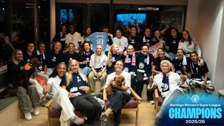 Group photo of Manchester City Women's team celebrating winning the Barclays Women's Super League 2025/26. The team is gathered in a room, some holding dogs, Manchester City scarves, and a jersey. Text signifies their championship