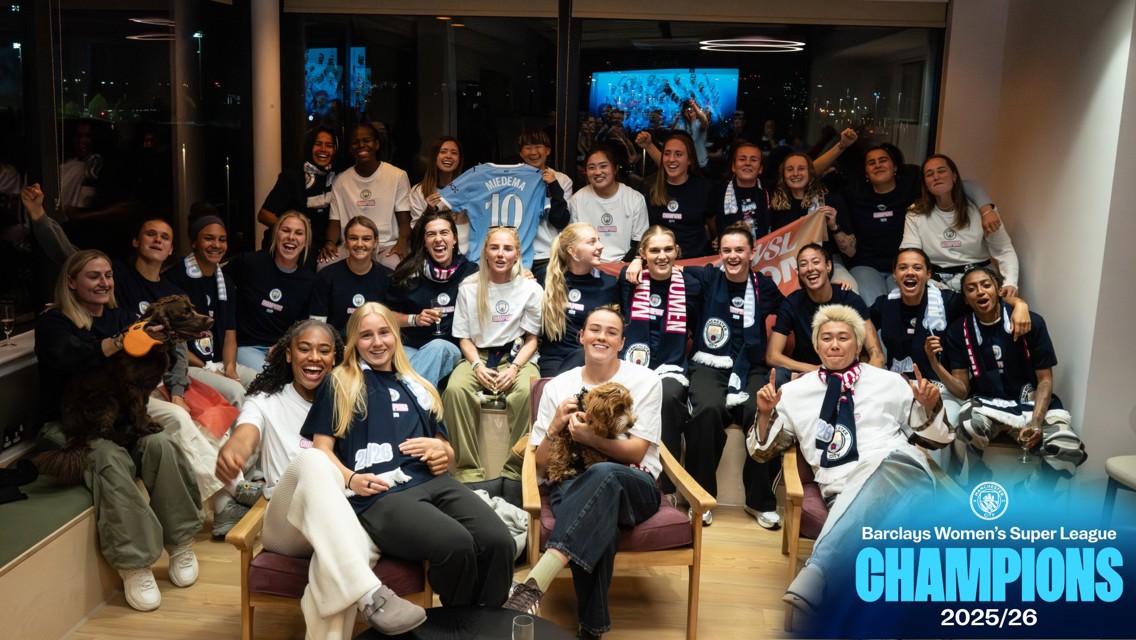 Group photo of Manchester City Women's team celebrating winning the Barclays Women's Super League 2025/26. The team is gathered in a room, some holding dogs, Manchester City scarves, and a jersey. Text signifies their championship