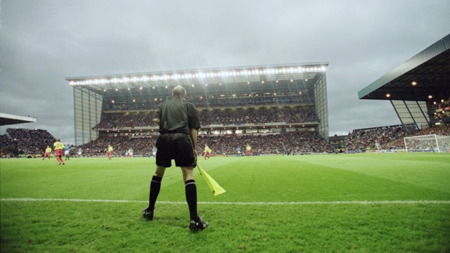A soccer match viewed from the sidelines with a referee in the foreground holding a flag. The stadium is filled with spectators under a cloudy sky.