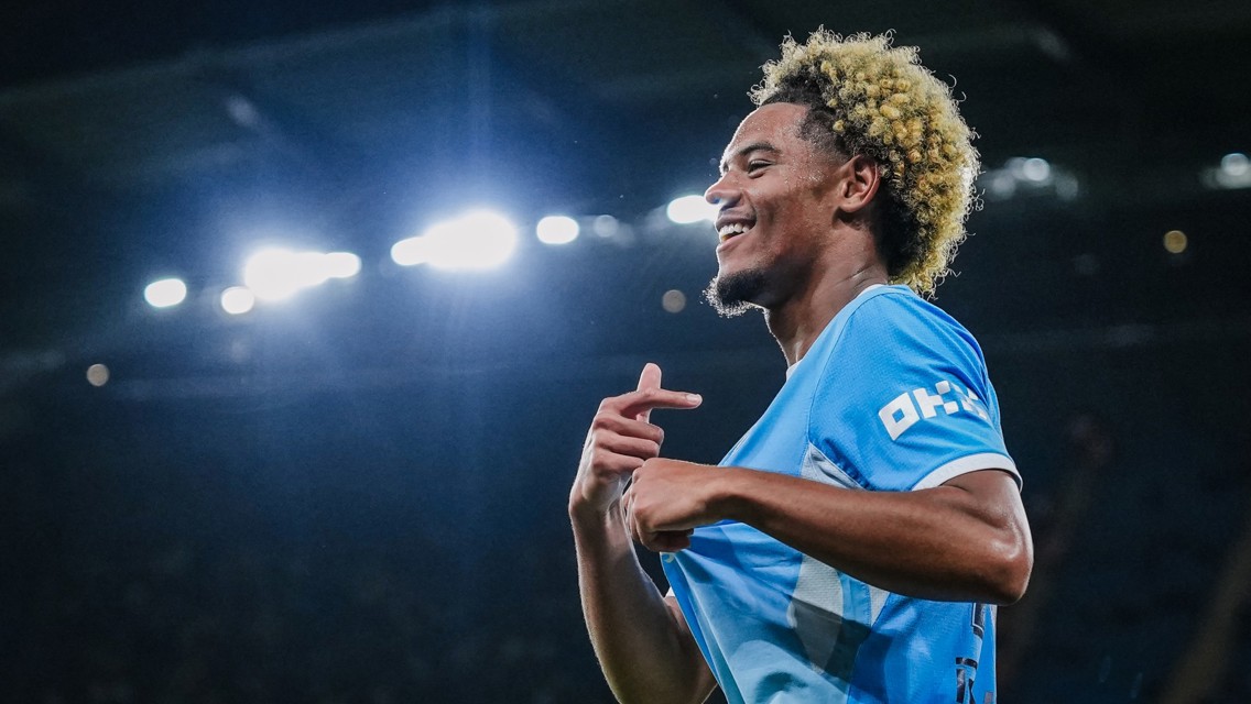 A soccer player in a light blue jersey is smiling and celebrating with bright stadium lights in the background.