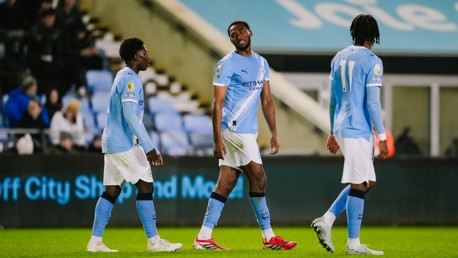 Three football players in Manchester City kits walking on a football field during a match, with the crowd in the background.
