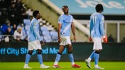 Three football players in Manchester City kits walking on a football field during a match, with the crowd in the background.