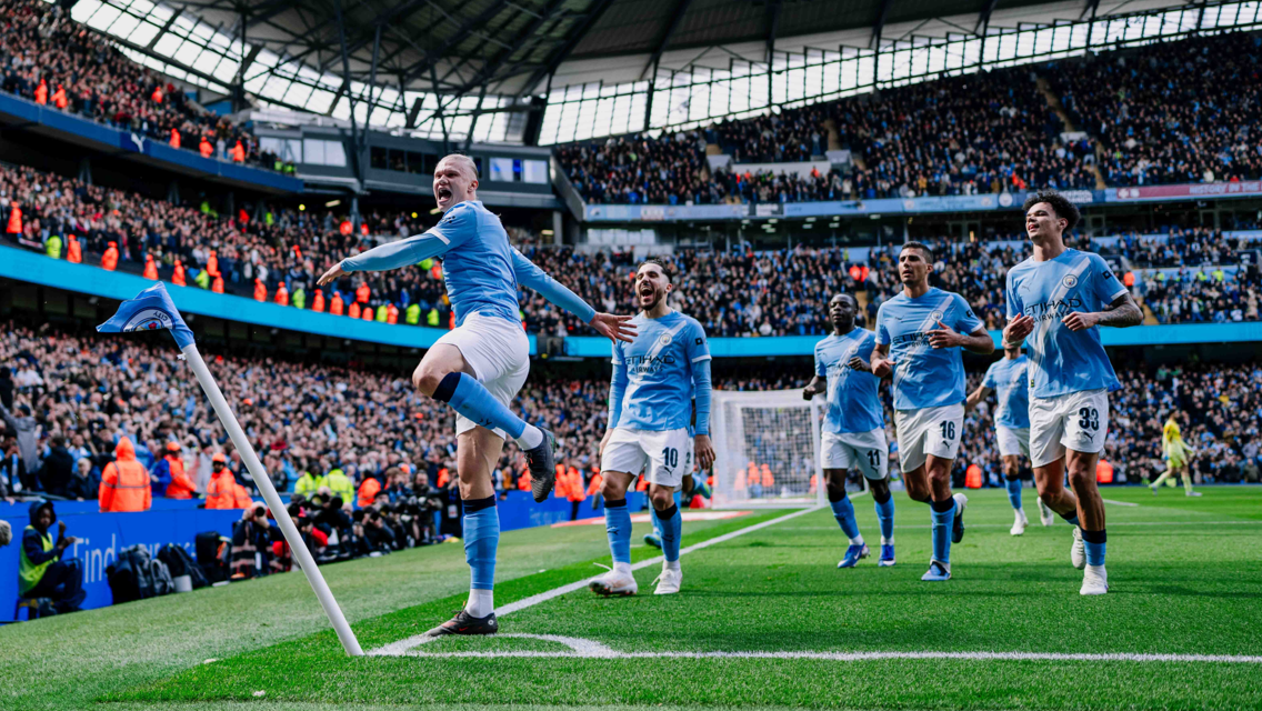 Soccer players in light blue jerseys celebrate near the corner flag at a packed stadium. The main player jumps with a raised knee, while others follow, running and smiling.