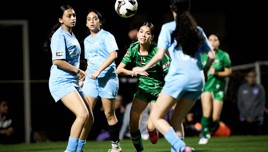 A women's soccer match with players in motion. They are wearing blue and green jerseys, and the ball is in the air.