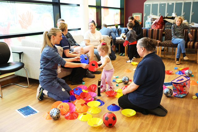 A community activity day with adults and children playing with colorful cones and balls on the floor. The setting appears to be a brightly lit room with seating and large windows. The faces of participants are blurred for privacy.