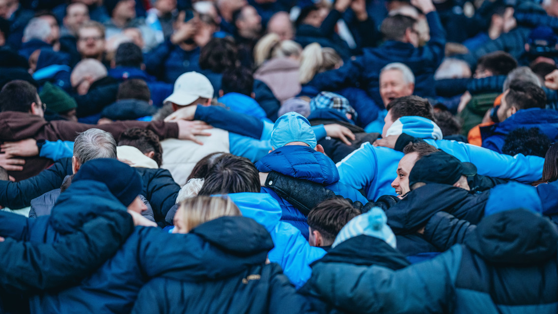 A large group of people huddled together in a stadium, with many wearing blue jackets and some with hats and scarves.