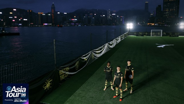 Three players in Manchester City jerseys stand on an illuminated football pitch next to a waterfront in Hong Kong as part of the Asia Tour '26.