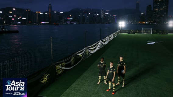 Three players in Manchester City jerseys stand on an illuminated football pitch next to a waterfront in Hong Kong as part of the Asia Tour '26.