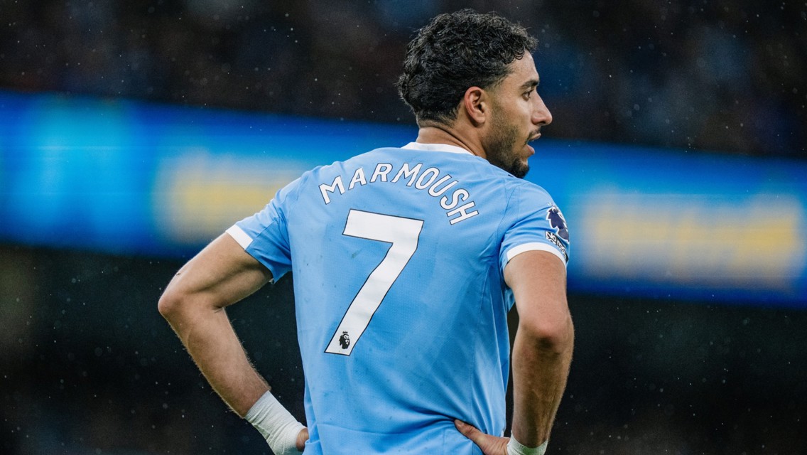 A soccer player wearing a blue jersey with 'Marmoush' and the number 7 on the back stands on the field during a match.
