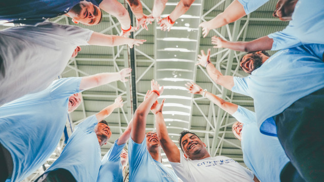 A group of people wearing blue shirts raising their arms in a team huddle under an indoor ceiling with bright lighting.