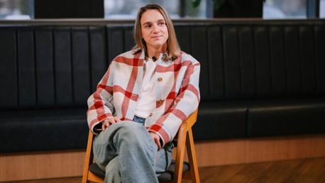 A woman wearing a red and white plaid jacket, white shirt, and jeans is sitting cross-legged on a wooden chair, with a dark bench in the background.