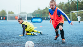 Children wearing sports bibs playing soccer on a blue outdoor field with goals visible in the background.