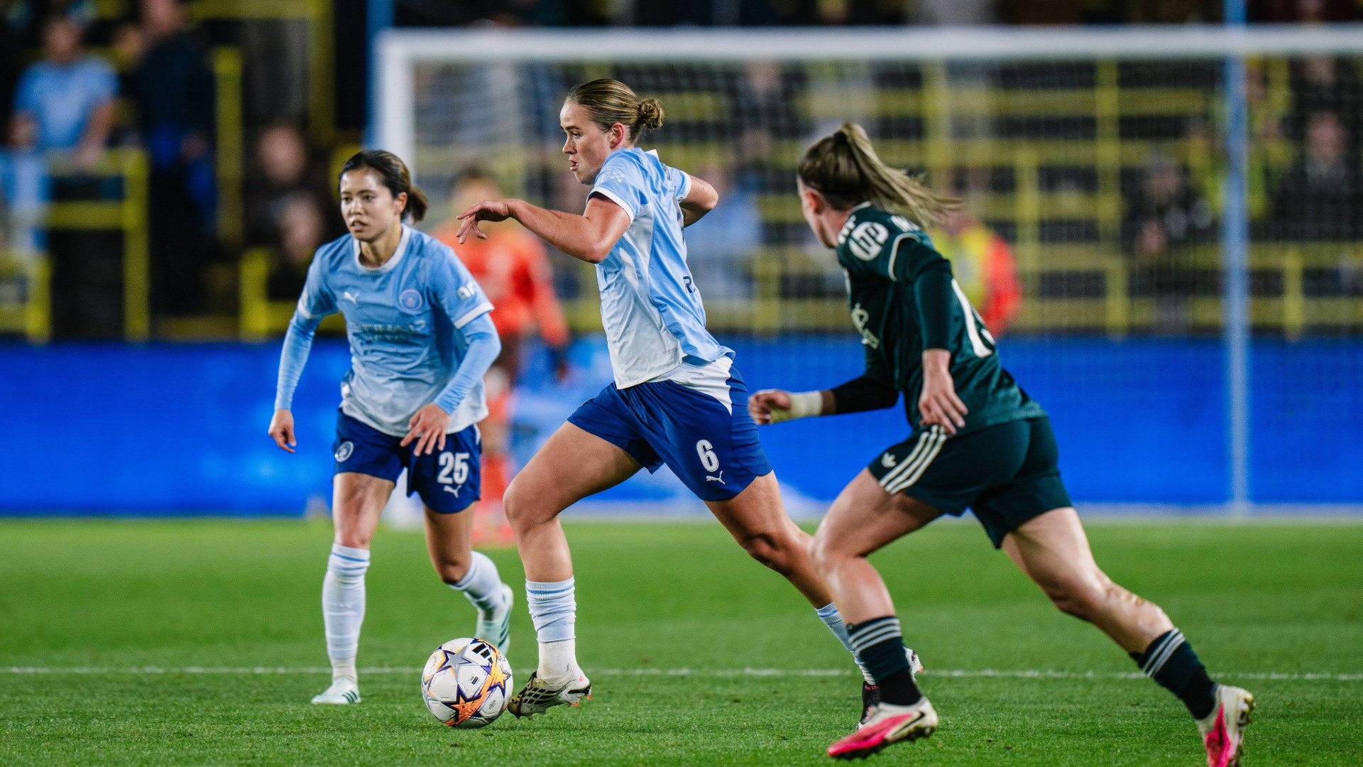 The image shows a women's soccer match with Manchester City players in light blue shirts competing against a player from the opposing team in dark green. The players are in action on the field focusing on the ball.