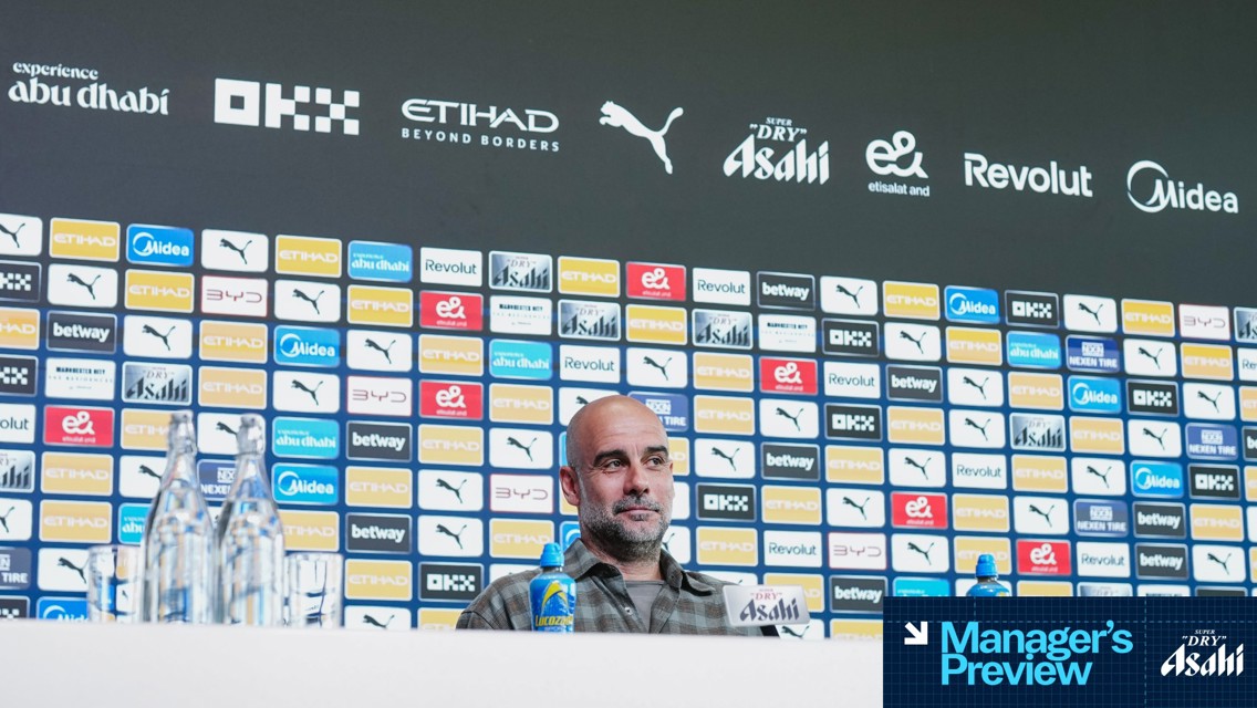 Press conference setup with branded backdrop and table items, including bottled drinks and a table sign showing Manager's Preview at Etihad Stadium.
