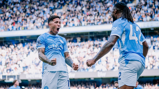 Two Manchester City players celebrating during a match at a stadium full of spectators, with one player wearing jersey number 42.
