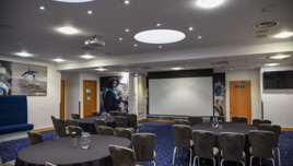 A conference room with round tables, chairs, and a projector screen. The room is decorated with sports-themed artwork on the walls.