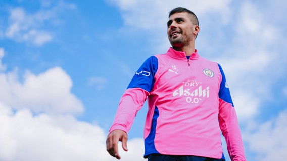 A person wearing a Manchester City training top, predominantly pink with blue sleeves and sponsors on the chest and arms. The sky is visible in the background.