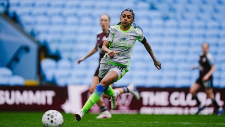 A soccer player in a gray and neon kit runs on a field with a ball nearby. The stadium has many empty blue seats.