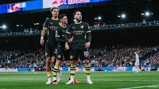 Three Manchester City players celebrating a goal in front of a crowd, wearing black and yellow kits with Etihad Airways sponsor logos, during a match against Leeds United.