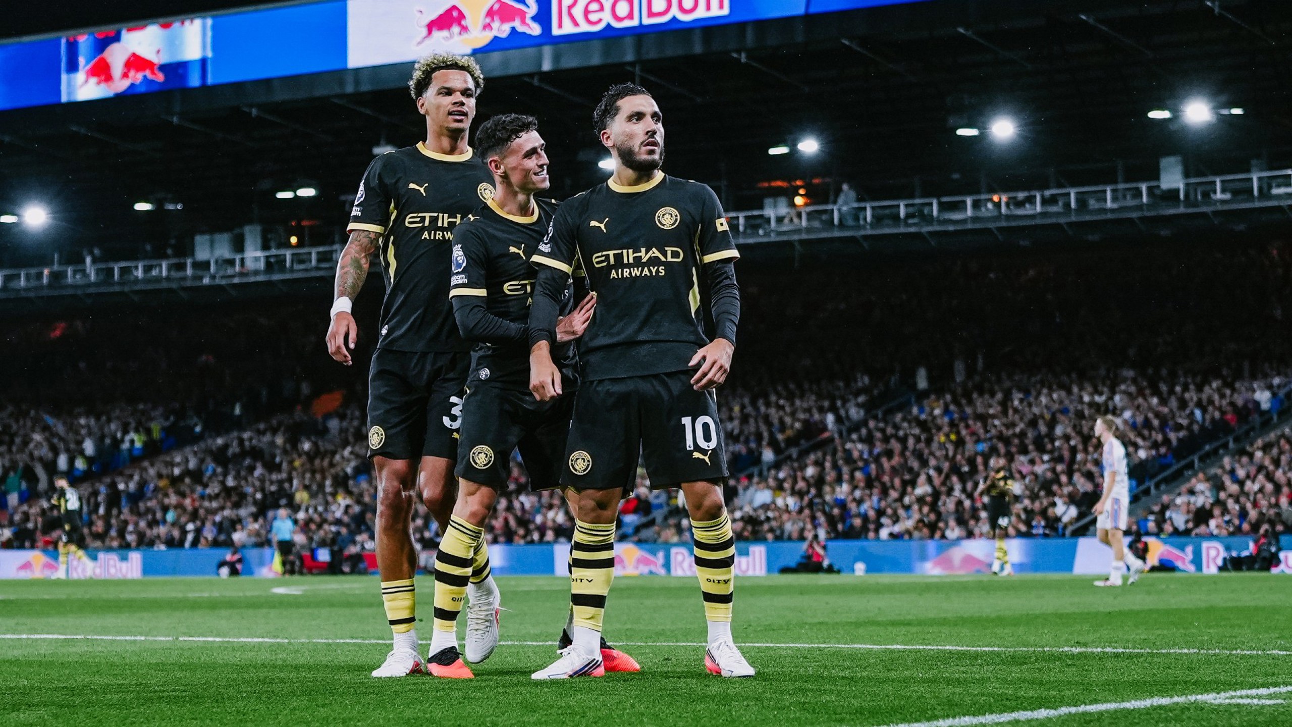 Three Manchester City players celebrating a goal in front of a crowd, wearing black and yellow kits with Etihad Airways sponsor logos, during a match against Leeds United.