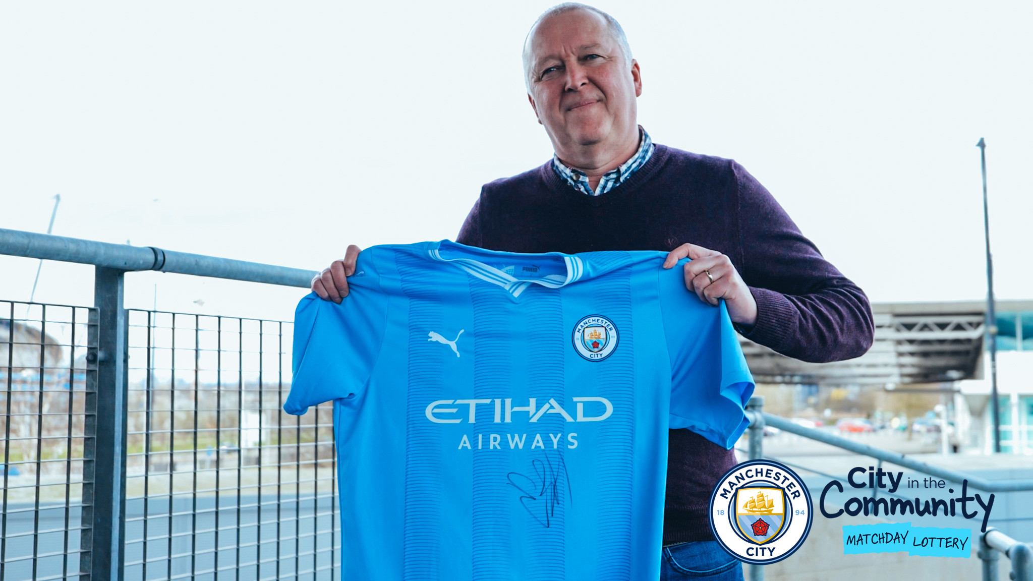 A man holding a signed Manchester City football jersey. The background includes a fence and an outdoor setting. The jersey features the Puma logo and Etihad Airways sponsor on the front.