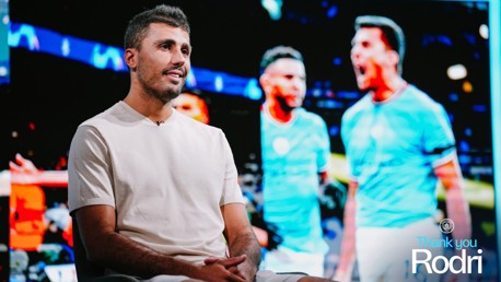 A person sitting and talking in front of a background showing two other people in Manchester City jerseys with the text 'Thank you Rodri' at the bottom.
