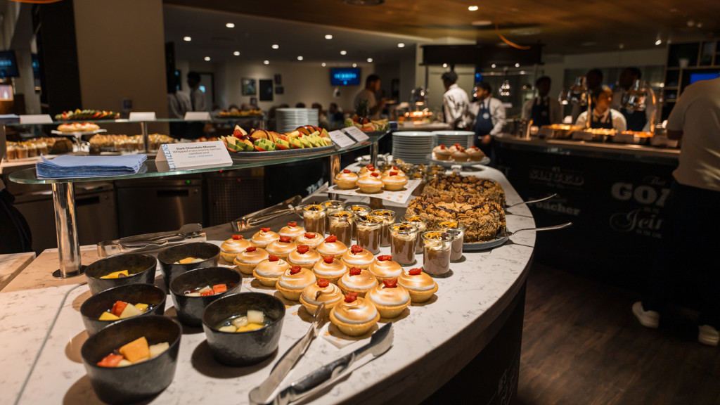 An assortment of desserts on display at a buffet, including fruit bowls, pastries, mousse cups, and cookies. Chefs and diners are visible in the background.