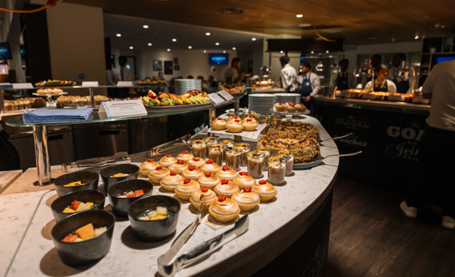 An assortment of desserts on display at a buffet, including fruit bowls, pastries, mousse cups, and cookies. Chefs and diners are visible in the background.
