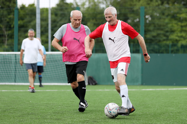 Two elderly men wearing Puma bibs play soccer on a field, one in a red and white outfit and one in a gray and black outfit. A soccer goal post is visible in the background.