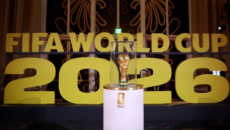 The image shows the FIFA World Cup trophy encased in glass in front of large yellow letters reading 'FIFA WORLD CUP 2026'.