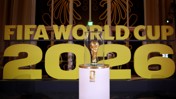 The image shows the FIFA World Cup trophy encased in glass in front of large yellow letters reading 'FIFA WORLD CUP 2026'.