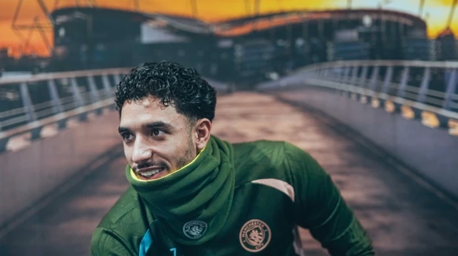 A Manchester City player in a green training kit with the club logo on it, standing on a bridge with the Etihad Stadium in the background during sunset.