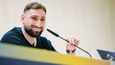 A person sitting at a table, speaking into a microphone. The person's face is blurred and they are wearing a dark sweater. A logo is partially visible in the foreground.