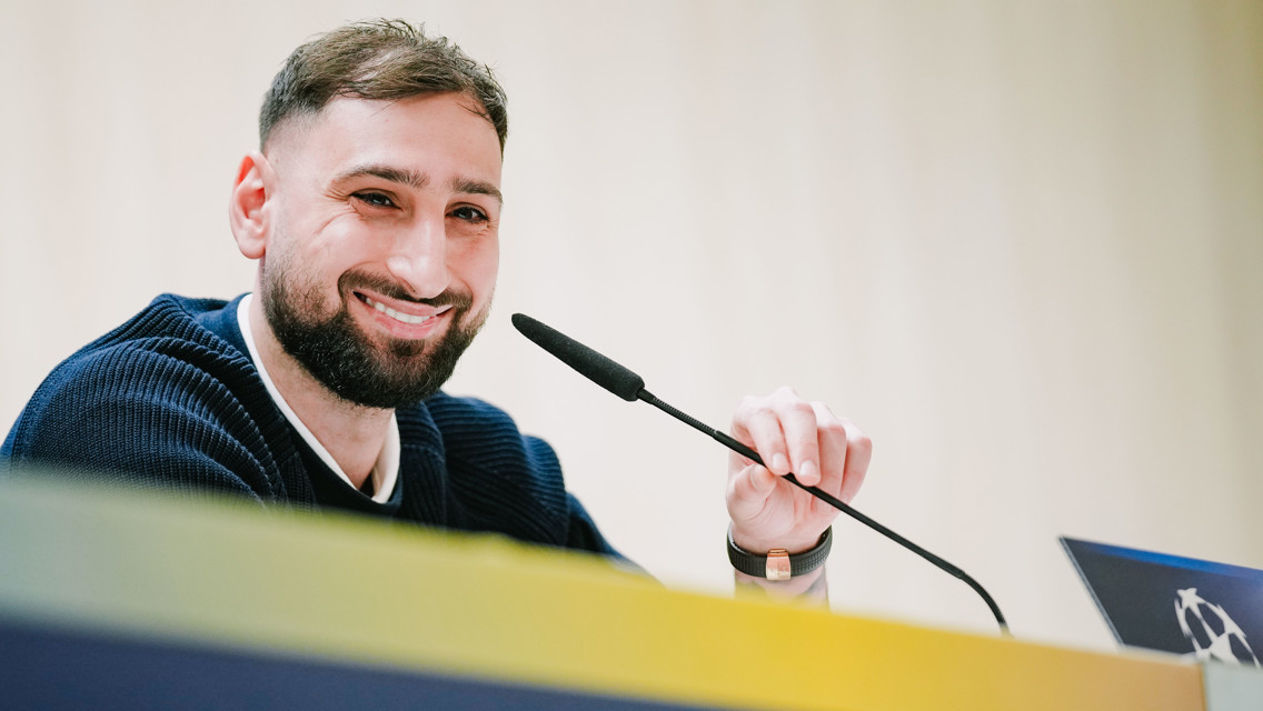 A person sitting at a table, speaking into a microphone. The person's face is blurred and they are wearing a dark sweater. A logo is partially visible in the foreground.