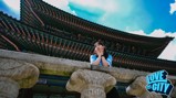 A woman in a blue shirt, standing at a stone railing, poses with a backdrop of traditional architecture. The image features a 'Love This City' logo in the corner.