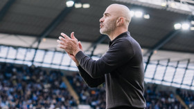 A bald man in a black jacket clapping on the sidelines of a football stadium.