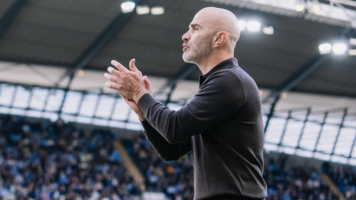 A bald man in a black jacket clapping on the sidelines of a football stadium.