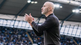 A bald man in a black jacket clapping on the sidelines of a football stadium.