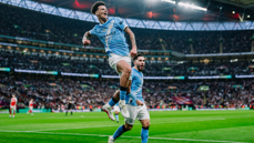 Two Manchester City players celebrate a goal during a match at a stadium with a large crowd.
