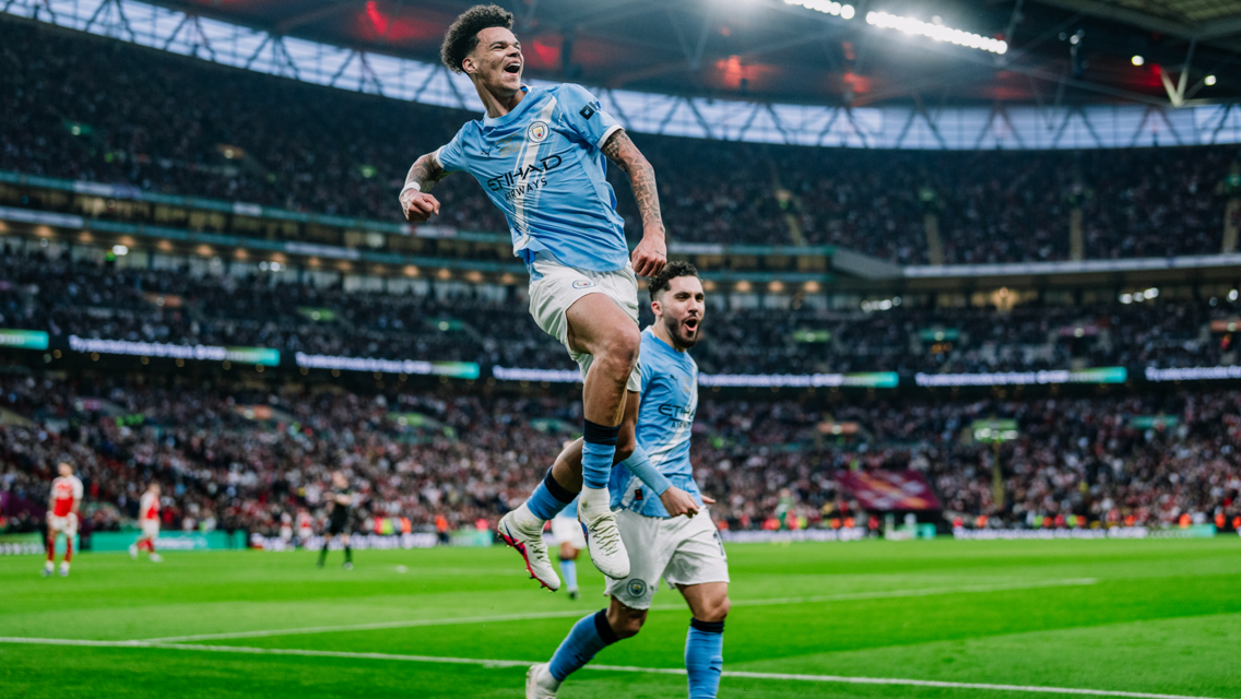 Two Manchester City players celebrate a goal during a match at a stadium with a large crowd.