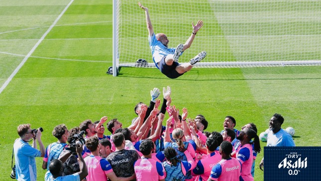 Football team in pink and blue uniforms celebrates by lifting a person in the air. Cameramen capture the moment.