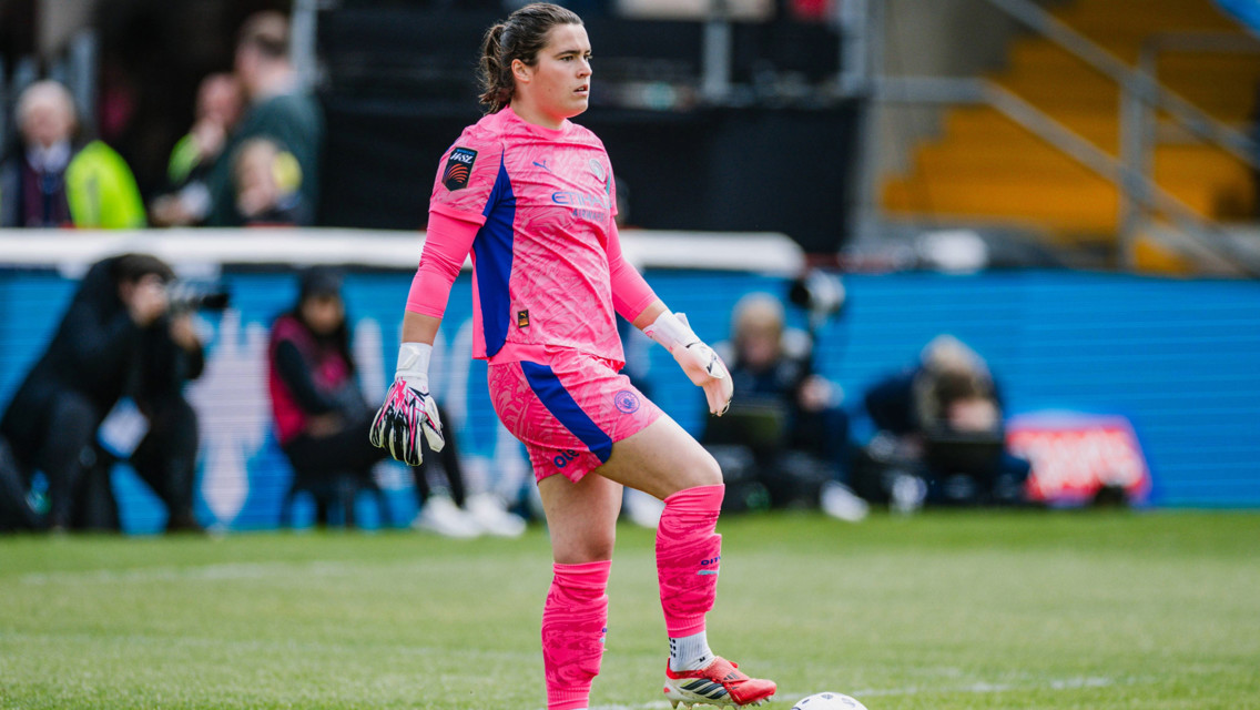 A soccer goalkeeper in pink kit preparing to kick a ball on a field, during a match.