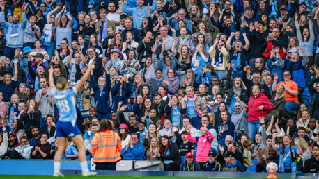 A crowd of fans at a stadium enthusiastically cheering and clapping, with a blurred player in the foreground raising their arms in celebration.
