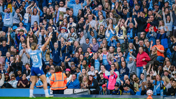 A crowd of fans at a stadium enthusiastically cheering and clapping, with a blurred player in the foreground raising their arms in celebration.