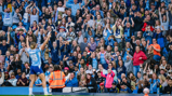 A crowd of fans at a stadium enthusiastically cheering and clapping, with a blurred player in the foreground raising their arms in celebration.