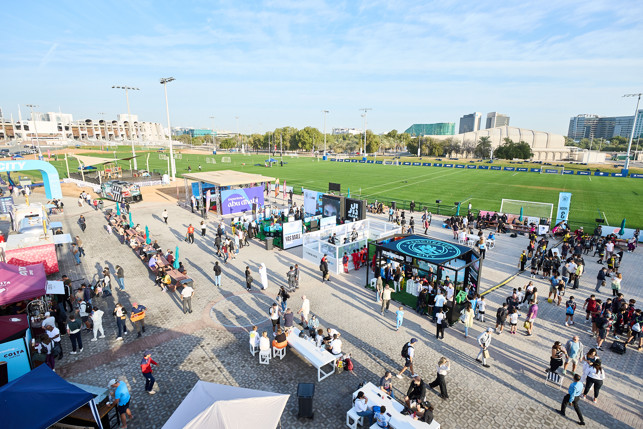 A busy outdoor event with many people walking around vendor stalls, soccer fields in the background, and a variety of promotional booths including one for Abu Dhabi. The sky is clear and the atmosphere is lively.